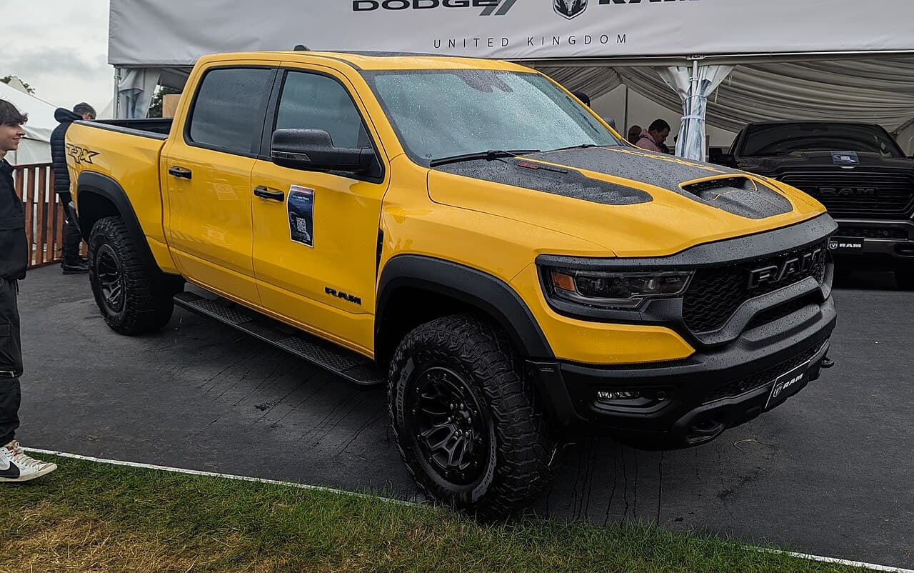 Front three-quarter view of a yellow Ram 1500 TRX under a 'Dodge // Ram United Kingdom' show tent, with a black Ram visible behind it.