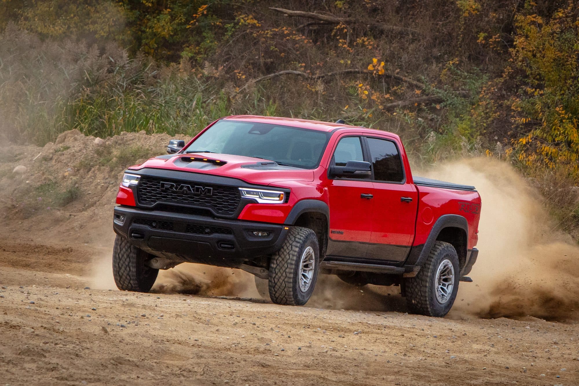 A red 2025 Ram 1500 RHO front three-quarter view, kicking up dust on a gravel track with the twin-turbo Hurricane hood scoop visible.