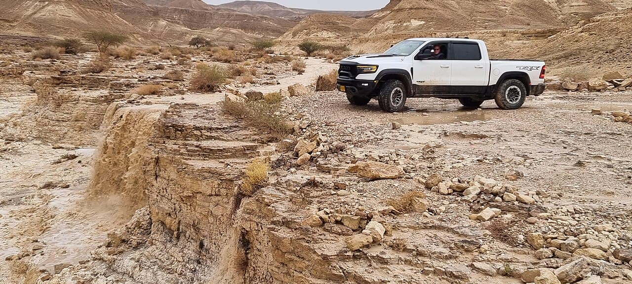 A white Ram 1500 TRX driving through a flash-flooded desert wadi (Nahal Tzin) in Israel's Arava valley, with water actively cascading over a rock ledge in the foreground.