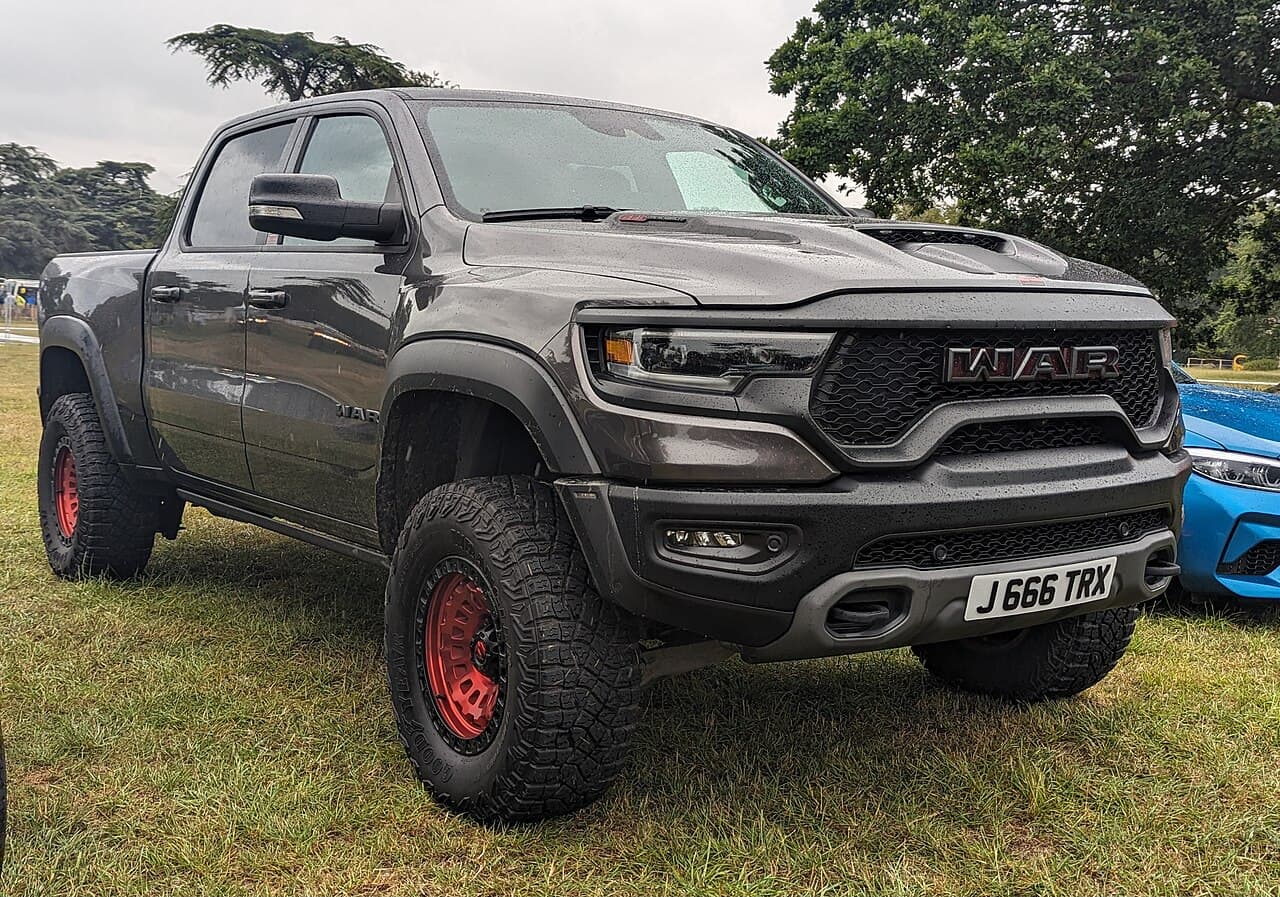 Front three-quarter view of a graphite-gray, rain-spattered Ram 1500 TRX on red wheels with a custom 'WAR' grille badge, at an outdoor UK event; the plate reads 'J 666 TRX'.
