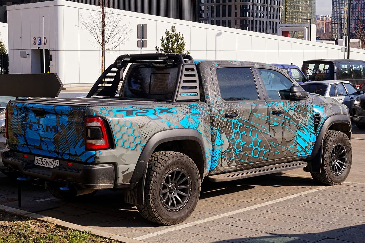 Rear three-quarter view of a Ram 1500 TRX wrapped in a gray/blue graphic livery with large 'TRX' lettering on the bed side and a roll cage with light bar, parked on a Moscow street.