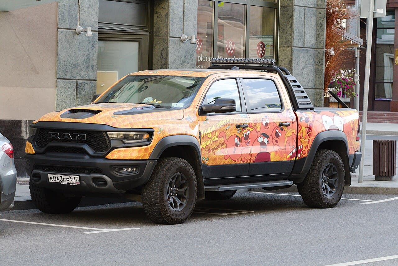 Side view of a Ram 1500 TRX wrapped in an orange honeycomb advertising livery, parked on a Moscow street with a light bar and roll cage fitted.