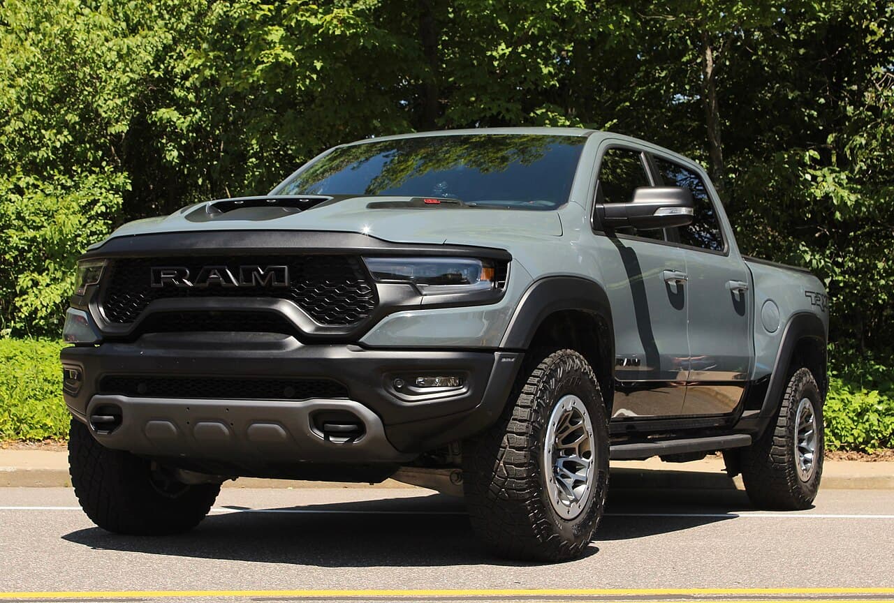 Front three-quarter view of a sage-gray Ram 1500 TRX parked roadside in front of trees.