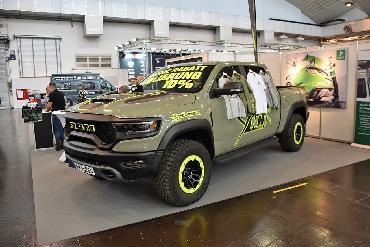 Front three-quarter view of a sage-green Ram 1500 TRX at an indoor promotional display with 'SPEED MONKEYS' branding and a clothing rack in the bed.
