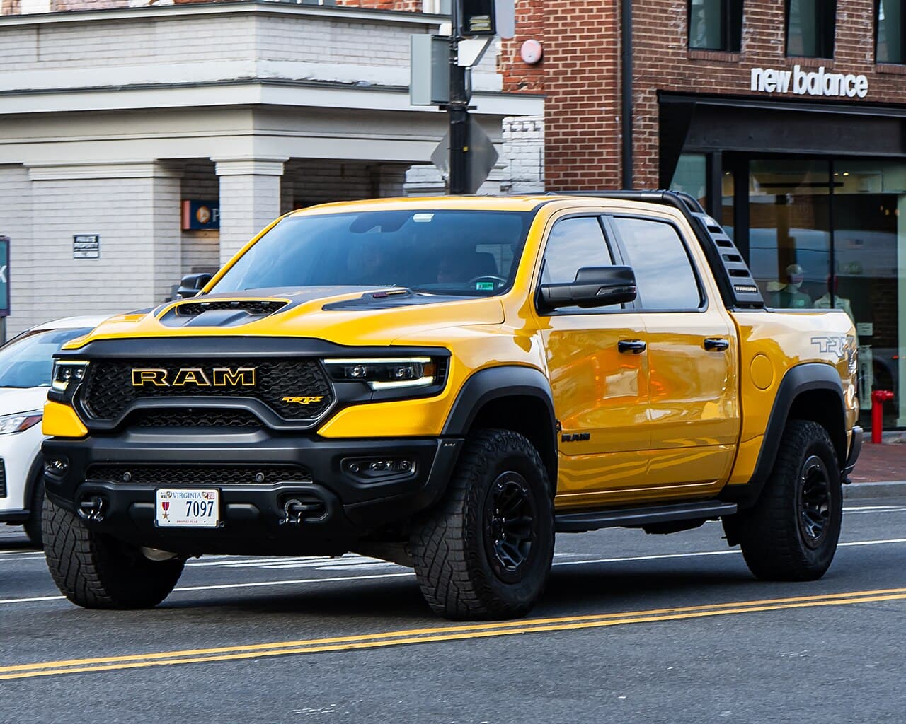 Front three-quarter view of a yellow Ram 1500 TRX driving down a city street in the Washington, DC metro area, past a New Balance storefront.