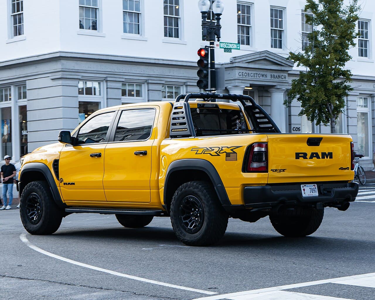 Rear three-quarter view of the same yellow Ram 1500 TRX turning through an intersection in Georgetown, Washington, DC, with a roll bar and light bar fitted.