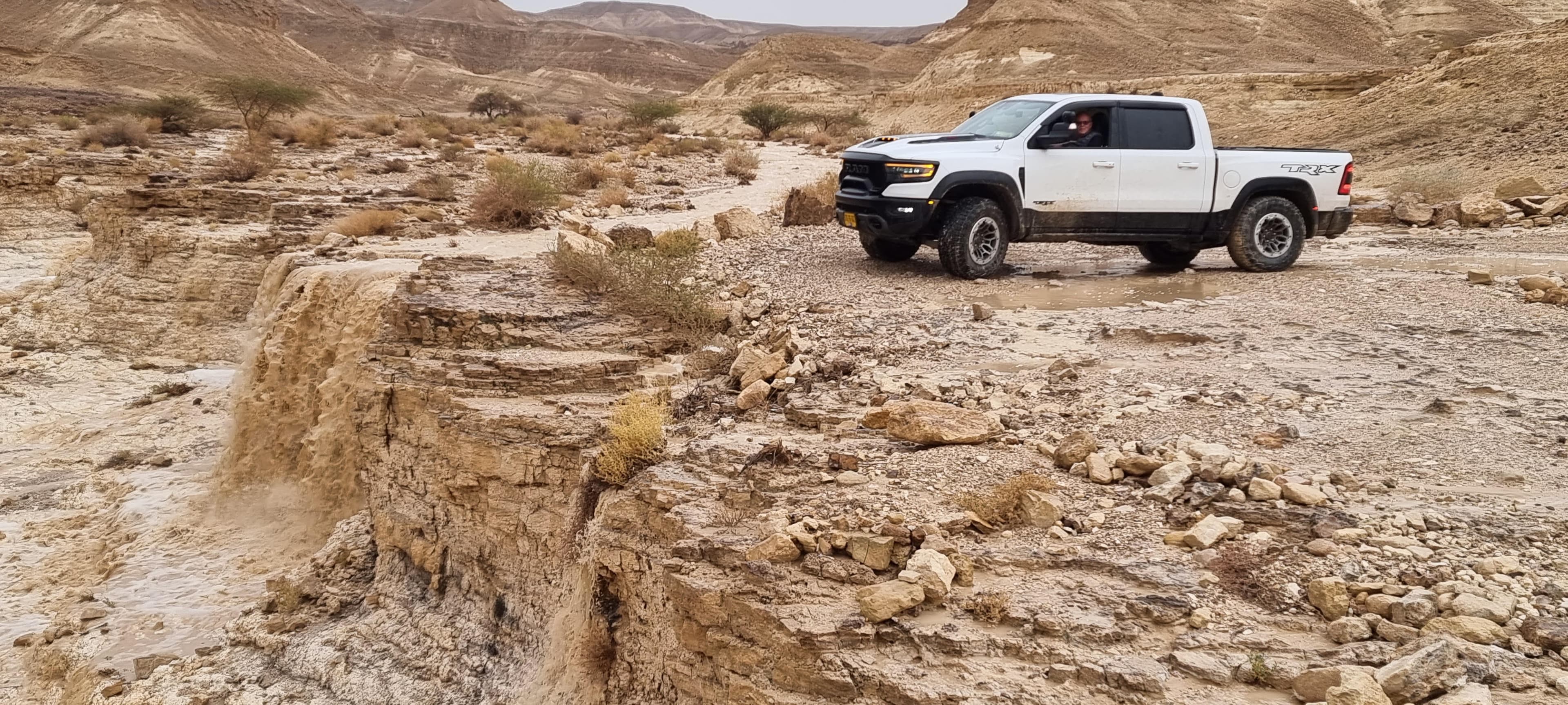 A white Ram 1500 TRX driving through a flash-flooded desert wadi (Nahal Tzin) in Israel's Arava valley, with water actively cascading over a rock ledge in the…